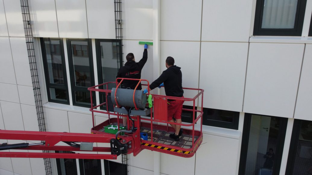 Zwei Glas- und Gebäudereiniger der Weber Gruppe im Einsatz. Auf einer Hubbühne bearbeiten sie eine Fassade am LANUV in Essen.