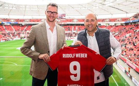 Andreas Weber (Geschäftsführer Rudolf Weber) und Fernando Carro de Prada (Vorsitzender Geschäftsführer Bayer Leverkusen) halten in der BayArena ein Trikot von Bayer Leverkusen in die Kamera.